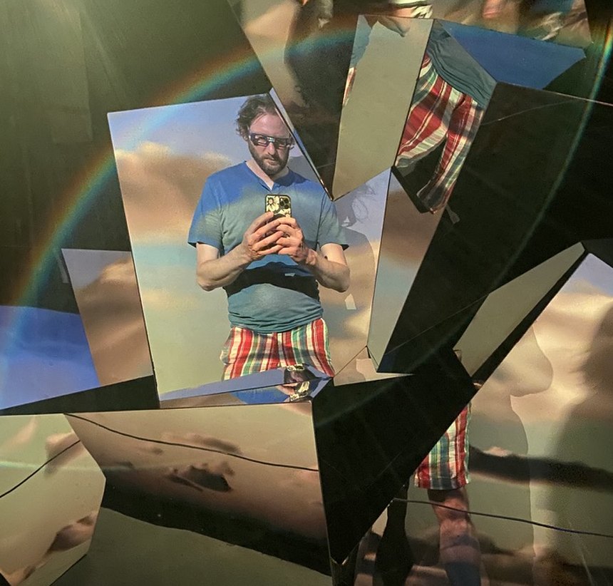 Eric Eaglstun taking a phone photo in a shattered-mirror art installation, a rainbow reflected across the glass shards